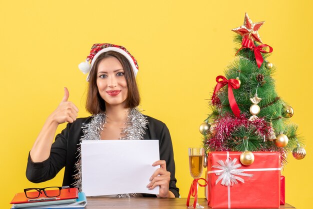 Lächelnde Geschäftsdame im Anzug mit Weihnachtsmannhut und Neujahrsdekorationen, die alleine arbeiten, machen ok Geste und sitzen an einem Tisch mit einem Weihnachtsbaum darauf im Büro