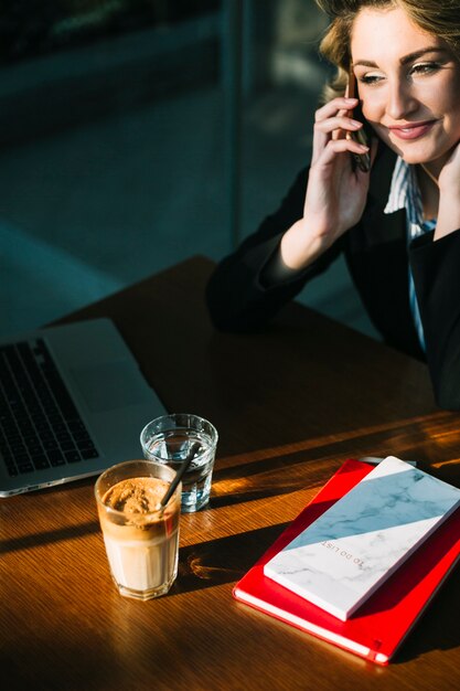 Lächelnde Geschäftsfrau, die auf Smartphone mit Laptop spricht; Schokoladenmilchshake und Bücher über Schreibtisch aus Holz