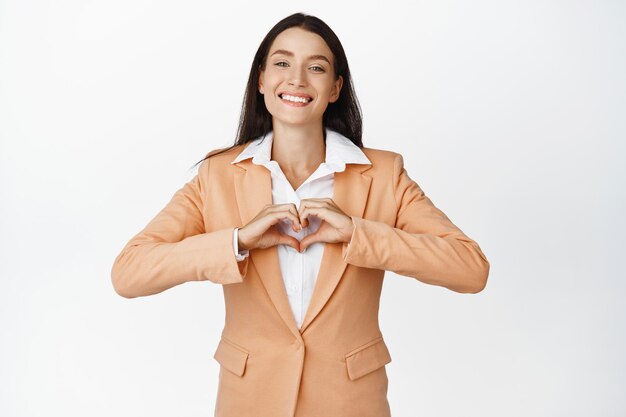 Lächelnde Geschäftsfrau, die Herzgeste zeigt, liebt ihre Kunden, die im Anzug vor weißem Hintergrund stehen