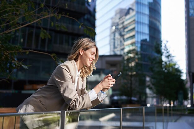 Lächelnde Geschäftsfrau, die ihr Handy in einer Pause außerhalb des Büros benutzt und auf Smartphone-Scre blickt