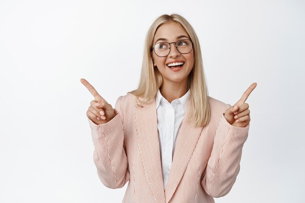 Lächelnde Geschäftsfrau, die mit dem Finger zur Seite zeigt und die Wahl zwischen zwei Optionen trifft, die in Anzug und Brille über weißem Hintergrund stehen