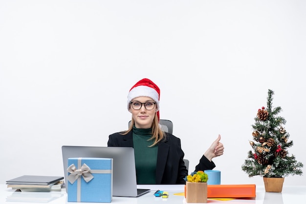 Lächelnde Geschäftsfrau mit einem Weihnachtsmannhut, der an einem Tisch mit einem Weihnachtsbaum und einem Geschenk darauf sitzt und etwas auf der linken Seite im Büro auf weißem Hintergrund zeigt