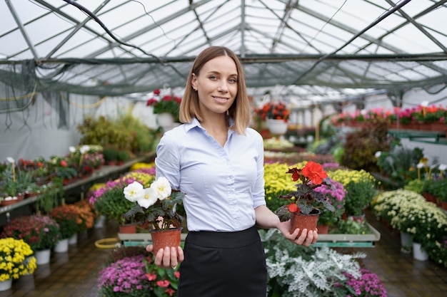 Lächelnde Geschäftsinhaberin in ihrem Kinderzimmer stehend, in Händen zwei Töpfe mit roten und weißen Blumen im Gewächshaus haltend