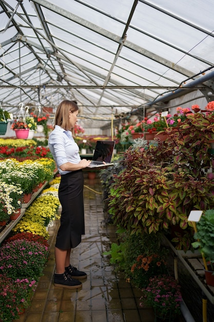 Lächelnde Gewächshausbesitzerin posiert mit einem Laptop in ihren Händen, die am Telefon mit vielen Blumen und Glasdach sprechen.