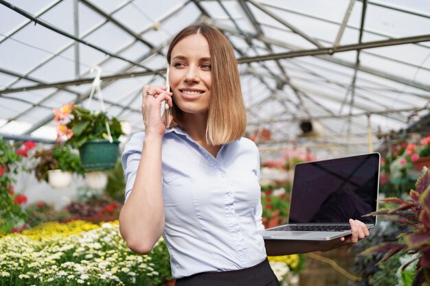 Lächelnde Gewächshausbesitzerin posiert mit einem Laptop in ihren Händen, die am Telefon mit vielen Blumen und Glasdach sprechen.