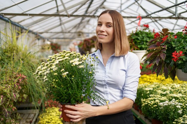 Lächelnde glückliche Floristin in ihrem Kinderzimmer, die Topfchrysanthemen in ihren Händen hält, während sie sich um die Gartenpflanzen im Gewächshaus kümmert