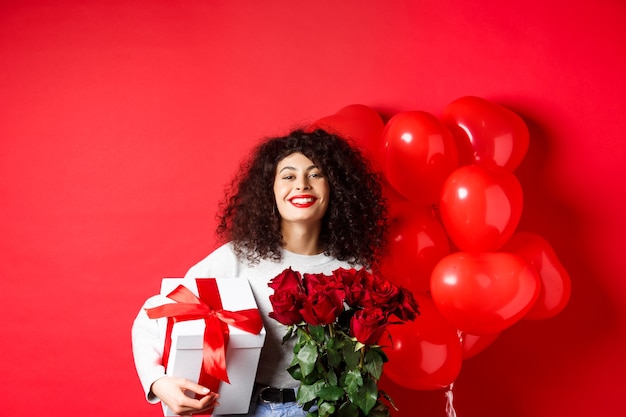 Lächelnde glückliche Frau, die eine Schachtel mit Geschenk und roten Rosen von ihrem Freund hält, der den Valentinstag feiert...