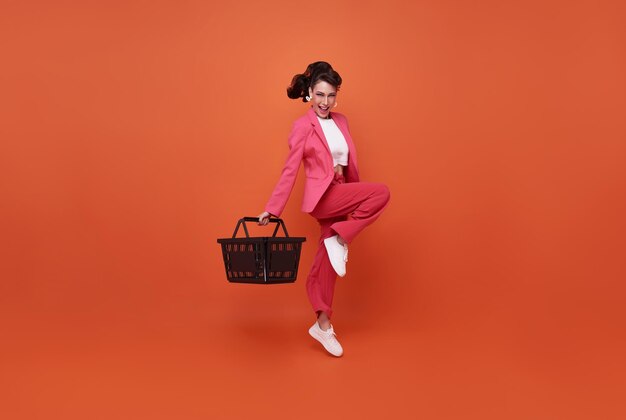Lächelnde glückliche Frau, die Einkaufskorb hält und auf orangefarbenem Hintergrund isoliert springt