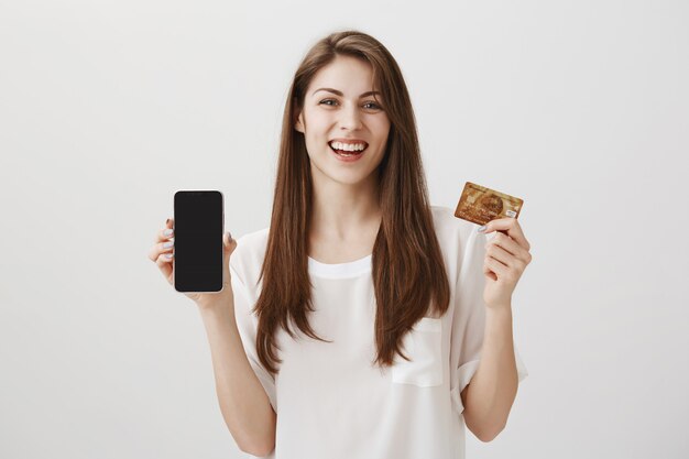 Lächelnde glückliche Frau, die Handyanzeige und Kreditkarte zeigt. Promo der Einkaufsanwendung