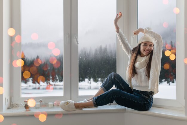 Lächelnde glückliche junge attraktive Frau in stilvollem weißem Strickpullover, Schal und Mütze, die zu Hause auf der Fensterbank zu Weihnachten sitzen und Spaß Händchenhalten, Winterwaldhintergrundansicht, Lichter bokeh haben