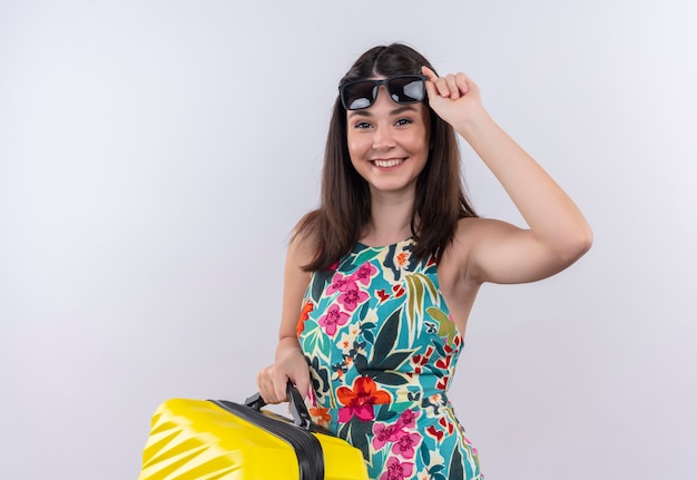 Lächelnde glückliche junge reisende Frau, die Koffer und Sonnenbrille auf isolierter weißer Wand hält