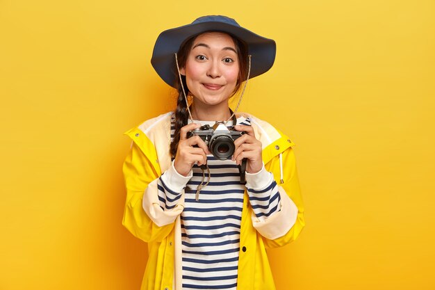 Lächelnde gut aussehende asiatische Frau mit langem Zopf, trägt Hut, gelben Regenmantel, hält Retro-Kamera, macht Fotos während ihrer fantastischen Reise, isoliert über gelber Wand