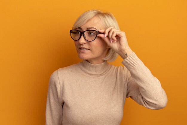 Lächelnde hübsche blonde slawische Frau in der optischen Brille schaut zur Seite auf Orange