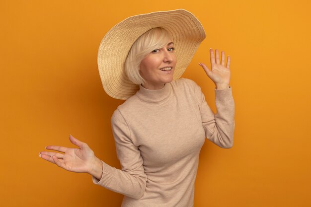 Lächelnde hübsche blonde slawische Frau mit Strandhut steht mit erhobenen Händen auf Orange