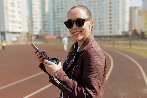 Lächelnde hübsche Dame mit dunkler Frisur in der Sonnenbrille unter Verwendung des Smartphones und des Musikhörens