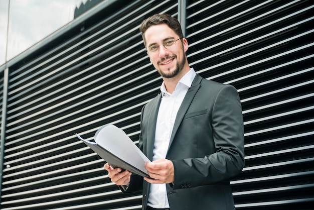 Lächelnde hübsche junge Geschäftsmannstellung außerhalb der Wand, die Ordner in der Hand hält