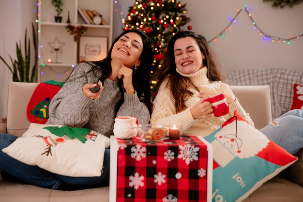 Lächelnde hübsche junge Mädchen halten TV-Fernbedienung und Tasse sitzen auf Sesseln und genießen die Weihnachtszeit zu Hause