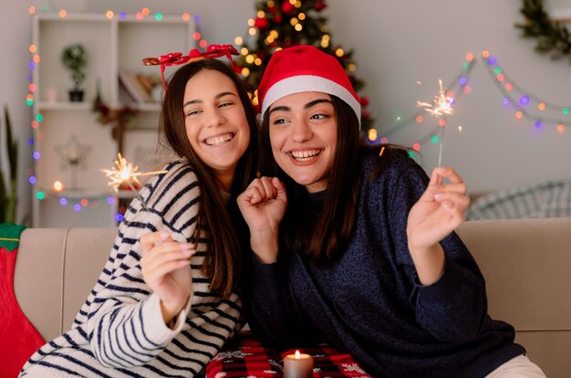 Lächelnde hübsche junge Mädchen mit Rentierbrille und Weihnachtsmütze, die Wunderkerzen halten und betrachten, die auf Sesseln sitzen und die Weihnachtszeit zu Hause genießen