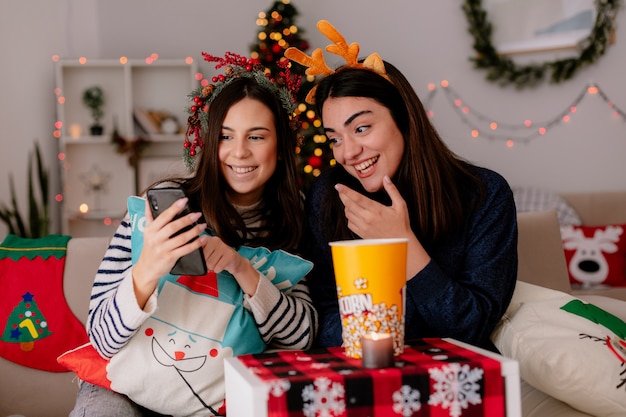 lächelnde hübsche junge Mädchen mit Stechpalmenkranz und Rentierstirnband betrachten Telefon, das auf Sesseln sitzt und Weihnachtszeit zu Hause genießt