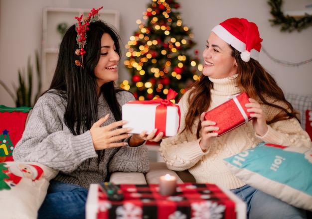 Lächelnde hübsche junge Mädchen mit Weihnachtsmütze und Stechpalmenkranz halten Geschenkboxen auf Sesseln sitzen und genießen die Weihnachtszeit zu Hause at