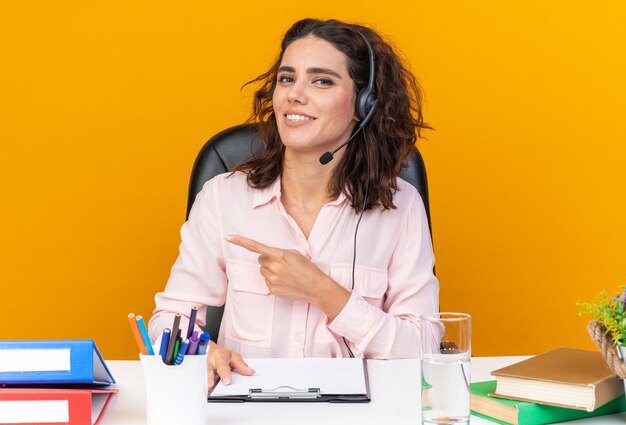 Lächelnde hübsche kaukasische Callcenter-Betreiberin auf Kopfhörern, die am Schreibtisch sitzen, mit Bürowerkzeugen, die auf die Seite zeigen, isoliert auf oranger Wand