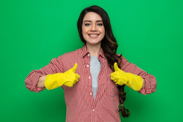 Lächelnde hübsche kaukasische Putzfrau mit Gummihandschuhen, die mit zwei Händen hochhält