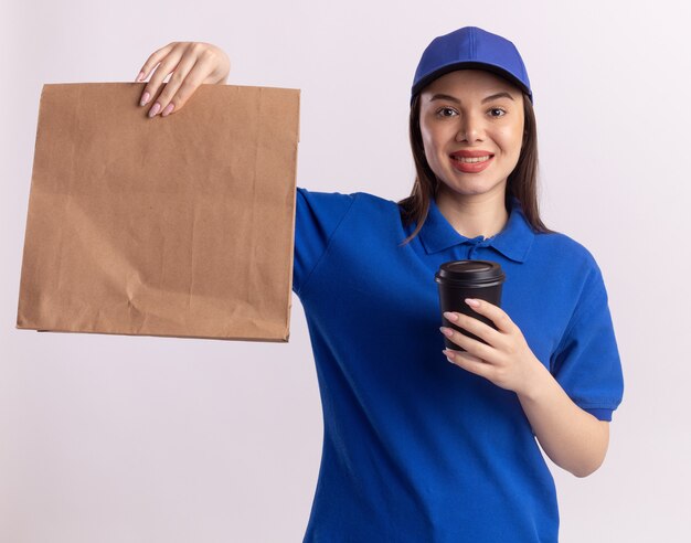 Lächelnde hübsche Lieferfrau in der Uniform hält Papierpaket und Pappbecher, die Kamera auf Weiß betrachten