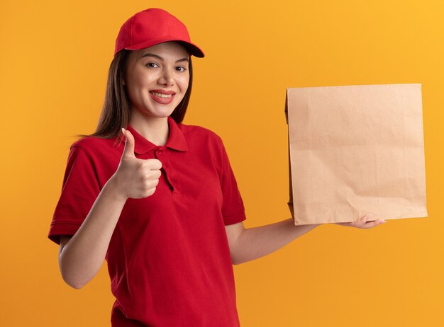 Lächelnde hübsche Lieferfrau in Uniform Daumen hoch und hält Papierpaket isoliert auf oranger Wand mit Kopierraum