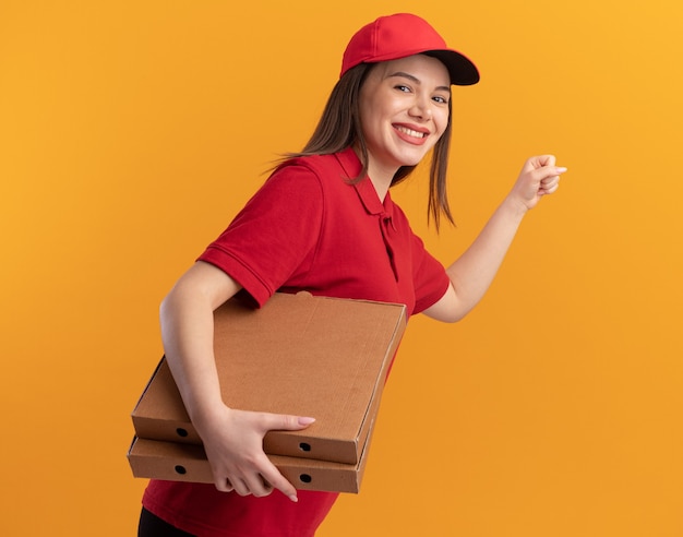 Lächelnde hübsche Lieferfrau in Uniform, die Faust hält und Pizzaschachteln auf Orange hält
