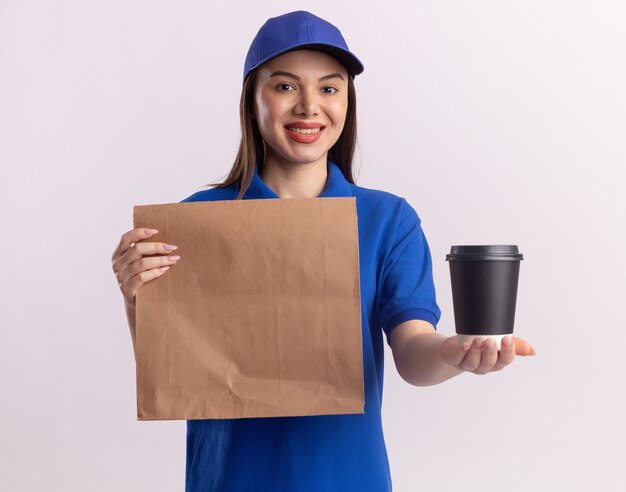 Lächelnde hübsche Lieferfrau in Uniform, die Papierpaket und Pappbecher lokalisiert auf weißer Wand mit Kopienraum hält