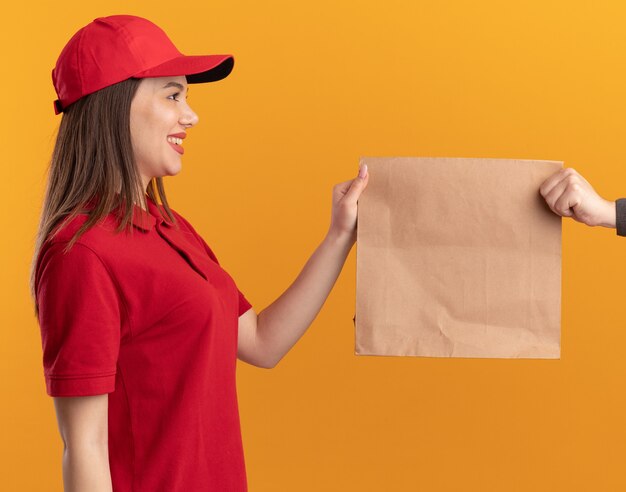 Lächelnde hübsche Lieferfrau in Uniform gibt jemandem, der auf einer orangefarbenen Wand mit Kopienraum isoliert ist, ein Papierpaket