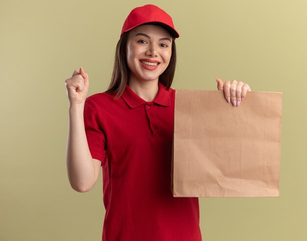 Lächelnde hübsche Lieferfrau in Uniform hält Faust und hält Papierpaket auf Olivgrün