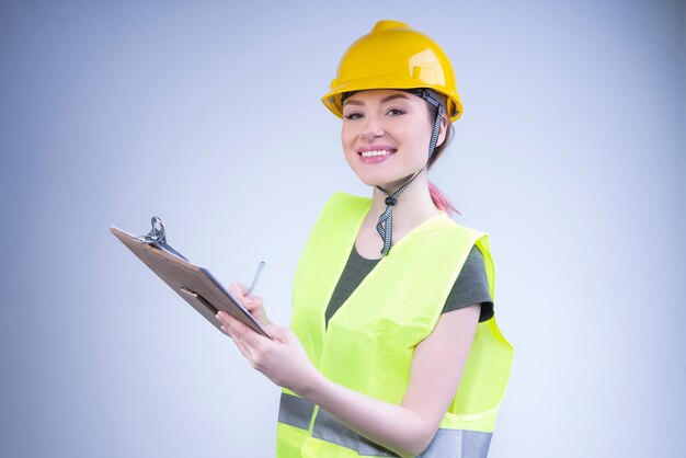 Lächelnde Ingenieurin in einem gelben Helm schreibt mit einem Stift in eine Zwischenablage