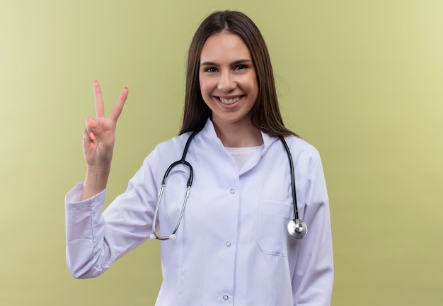 Lächelnde junge Ärztin, die das medizinische Kleid des Stethoskops trägt, das Friedensgeste auf grünem Hintergrund zeigt