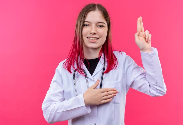 lächelnde junge Ärztin, die das medizinische Kleid des Stethoskops zeigt, das Pistolengeste zeigt, legte ihre Hand auf Herz auf rosa isolierte Wand