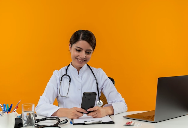 Lächelnde junge ärztin, die medizinische robe und stethoskop trägt, sitzt am schreibtisch mit medizinischen werkzeugen und laptop unter verwendung des mobiltelefons lokalisiert auf gelber wand