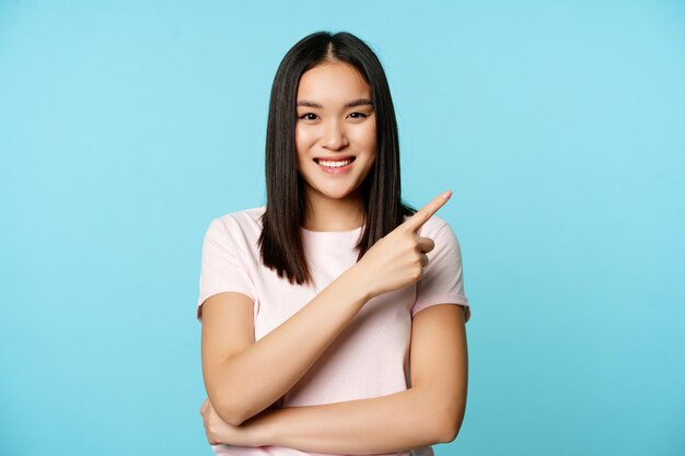 Lächelnde junge Asiatin 20 Jahre alt, zeigt mit dem Finger auf die obere rechte Ecke und zeigt Promo-Banner, blauer Hintergrund