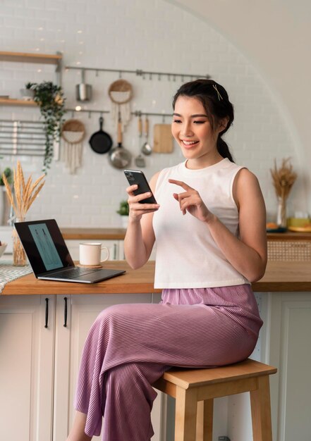 Lächelnde junge asiatin, die handy benutzt, während sie zu hause mit laptop in der küche sitzt