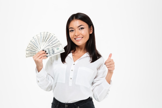 Lächelnde junge asiatische Frau, die Geld hält, das Daumen hoch zeigt.
