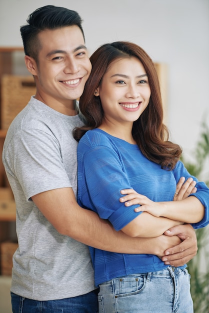 Lächelnde junge asiatische Paare, die zuhause stehen und umarmen