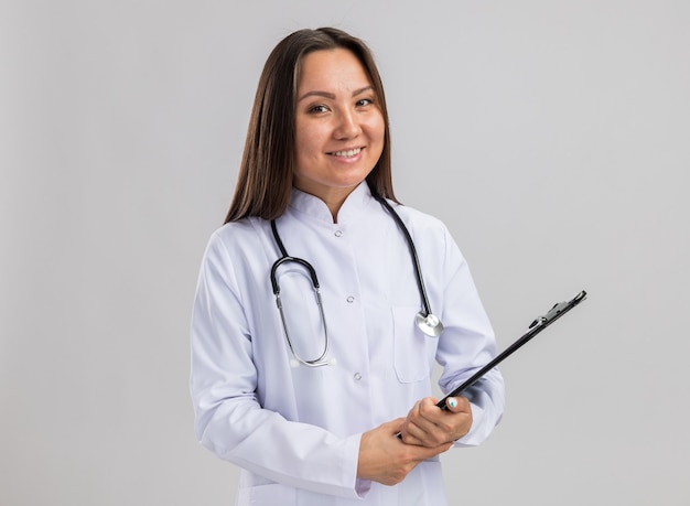Lächelnde junge asiatische Ärztin mit medizinischem Gewand und Stethoskop mit Zwischenablage und Blick in die Kamera isoliert auf weißer Wand