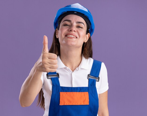 Lächelnde junge Baumeisterin in Uniform, die Daumen oben auf lila Wand lokalisiert zeigt