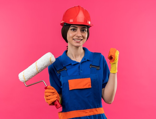 Lächelnde junge Baumeisterin in Uniform mit Handschuhen, die eine Walzenbürste hält und die Ja-Geste einzeln auf rosa Wand zeigt