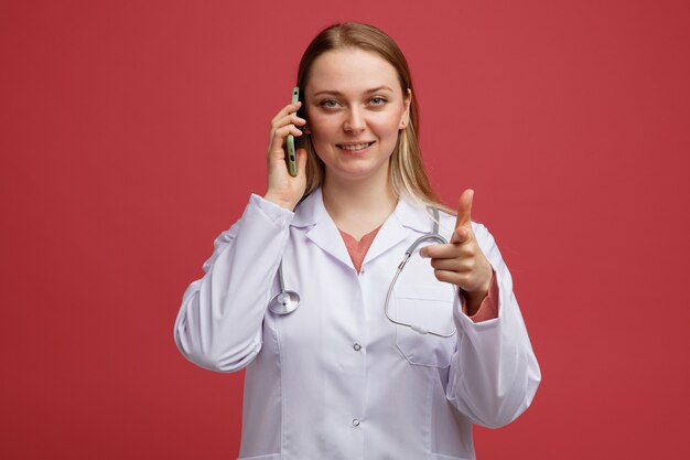 Lächelnde junge blonde Ärztin, die medizinische Robe und Stethoskop um den Hals trägt und am Telefon spricht und auf Kamera zeigt