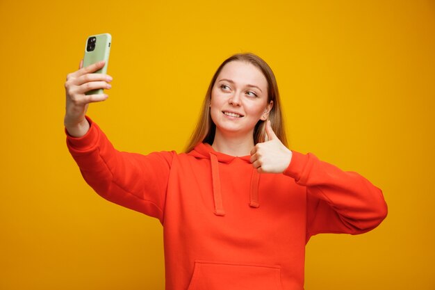Lächelnde junge blonde Frau, die selfie zeigt Daumen zeigt