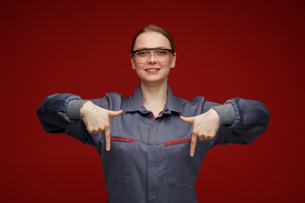 Kostenloses Foto lächelnde junge blonde ingenieurin, die uniform und schutzbrille trägt, zeigt nach unten