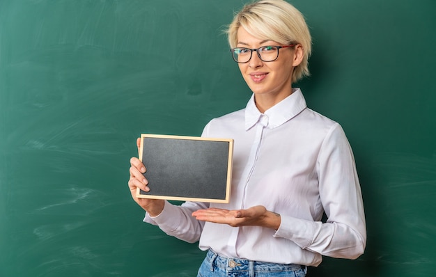 Kostenloses Foto lächelnde junge blonde lehrerin mit brille im klassenzimmer, die vor der tafel steht und eine mini-tafel zeigt, die nach vorne mit kopienraum schaut