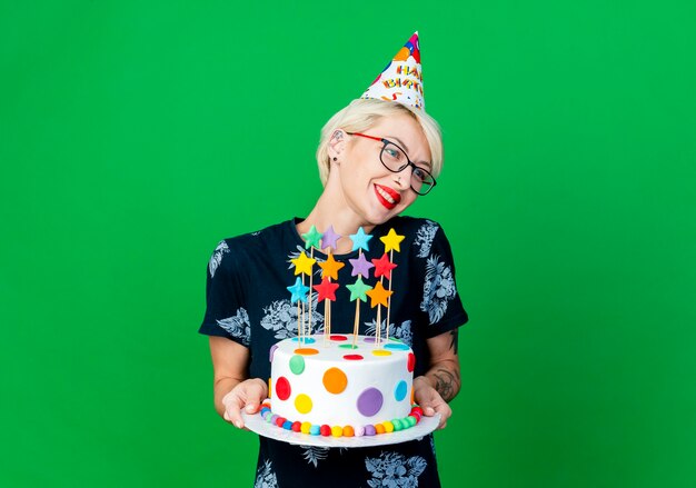 Lächelnde junge blonde Partygirl, die Brille und Geburtstagskappe hält, die Geburtstagstorte mit Sternen hält, die Seite lokalisiert auf grünem Hintergrund mit Kopienraum betrachten