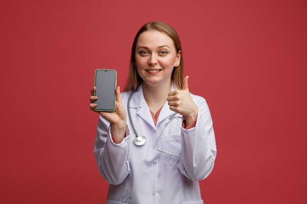 Lächelnde junge blonde Ärztin, die medizinische Robe und Stethoskop um den Hals trägt, hält Handy, das Daumen oben zeigt