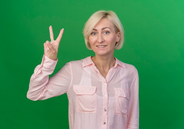 Lächelnde junge blonde slawische Frau, die Friedenszeichen lokalisiert auf grünem Hintergrund mit Kopienraum tut